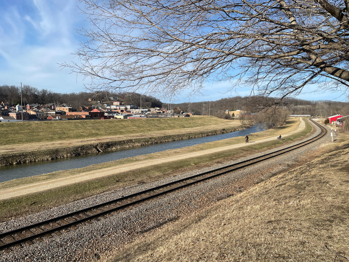 Scenic landscape in Grant Park, Galena, Illinois showing a curving railway beside a narrow river, two cyclists riding along a dirt trail, and the town’s historic buildings in the background on a clear early‑spring day.