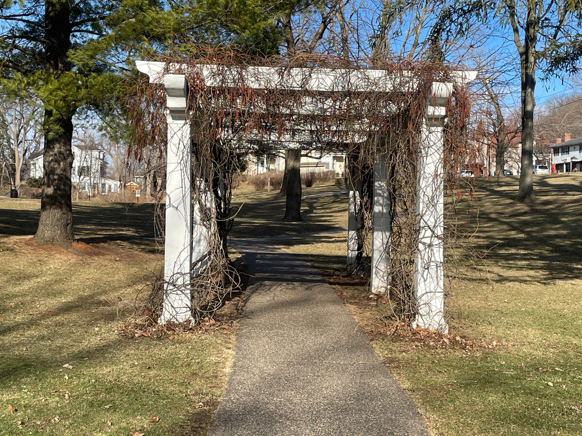 White pergola with dormant vines over a paved walkway in Grant Park, Galena, Illinois, surrounded by leafless winter trees and nearby homes.
