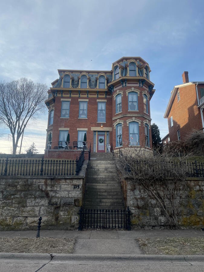 A tall Second Empire–style brick home on a hillside in Galena, Illinois, featuring a mansard roof, arched windows, decorative trim, a stone retaining wall, and a wrought‑iron gate with a leafless tree in front under a partly cloudy sky.