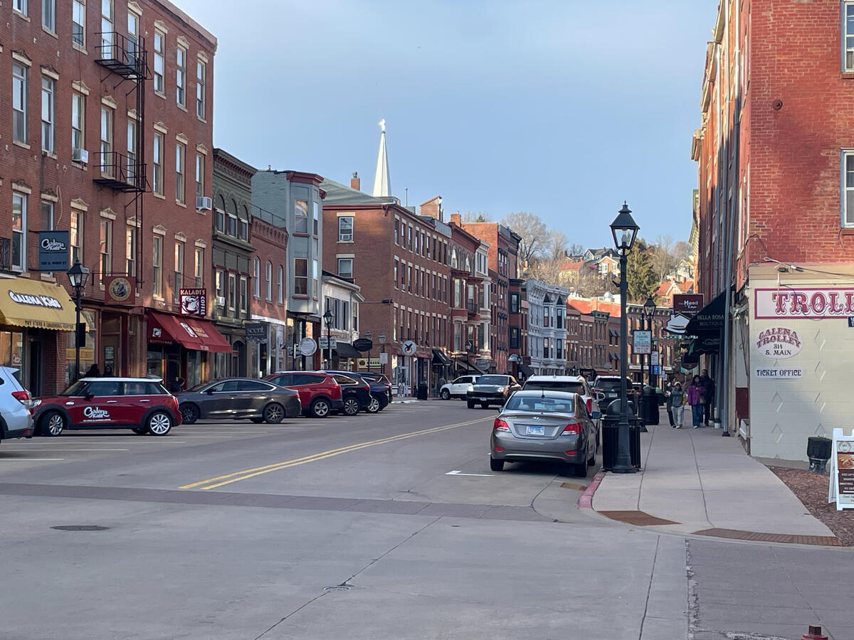Historic Main Street in downtown Galena, Illinois, featuring 19th‑century brick storefronts, parked cars, pedestrians, and the Galena Trolley Company ticket office with a church steeple rising in the background.