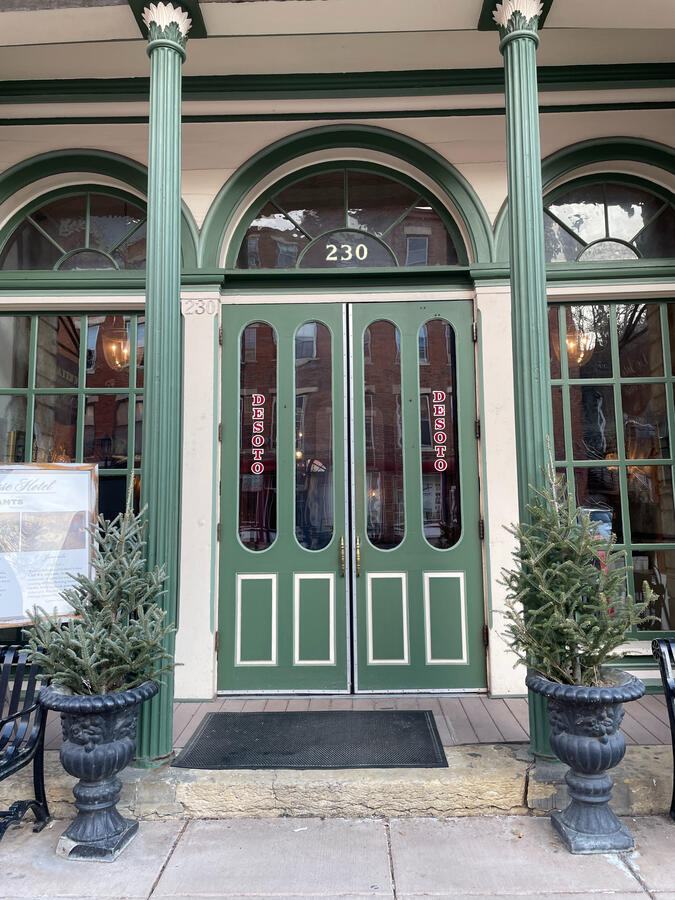 Entrance to the DeSoto House Hotel in Galena, Illinois, with green double doors featuring vertical red “DeSoto” lettering on both sides, an arched transom window displaying the number 230, evergreen planters, and a bench and signboard along the sidewalk in