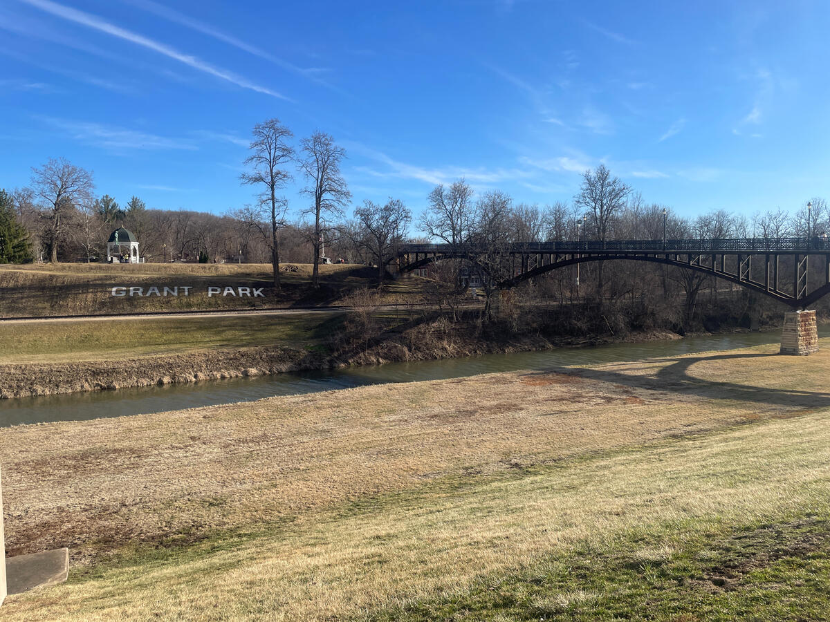 View of the arched metal bridge crossing the Galena River in Grant Park, Galena, Illinois, with “Grant Park” spelled out on the hillside, leafless trees, and a small green-roof gazebo under a clear blue sky.