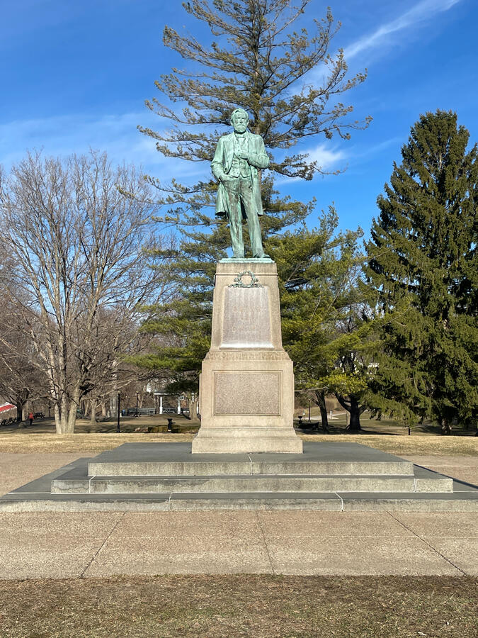 Bronze statue of Ulysses S. Grant on a tall stone pedestal in Grant Park, Galena, Illinois, surrounded by paved walkways, grass, and early‑spring trees under a clear blue sky.