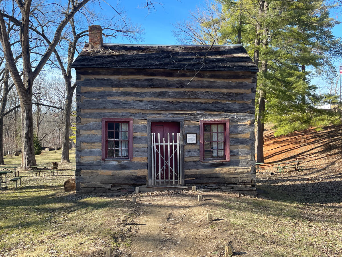 Historic log cabin in Galena, Illinois, featuring hand‑hewn timber walls, red‑trimmed windows, a brick chimney, and a small white gate, set in a wooded park with leafless trees and picnic tables in early spring.
