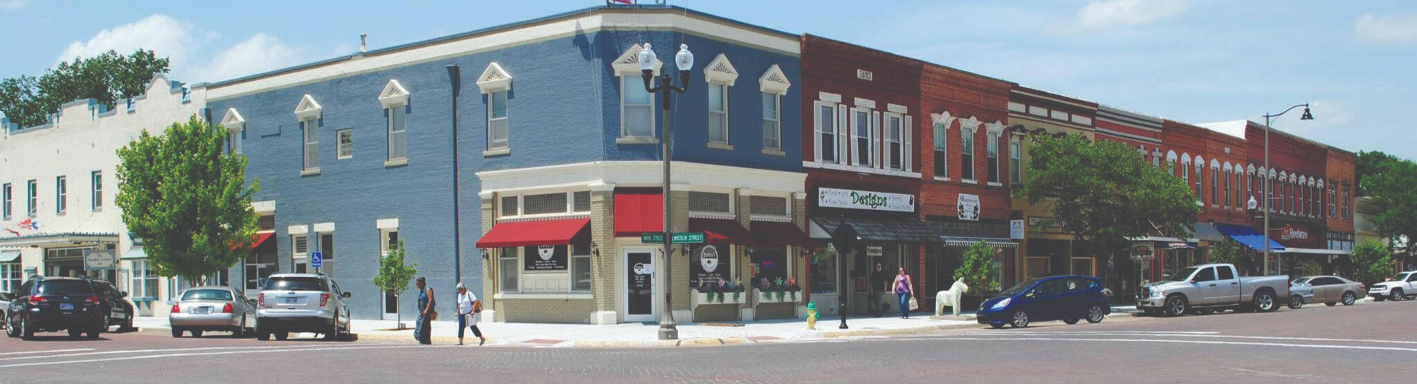 lindsborg-kansas-downtown-intersection-sunny-day A sunny downtown intersection in Lindsborg, Kansas, with historic brick buildings, storefronts, parked cars, and people walking along the sidewalk.