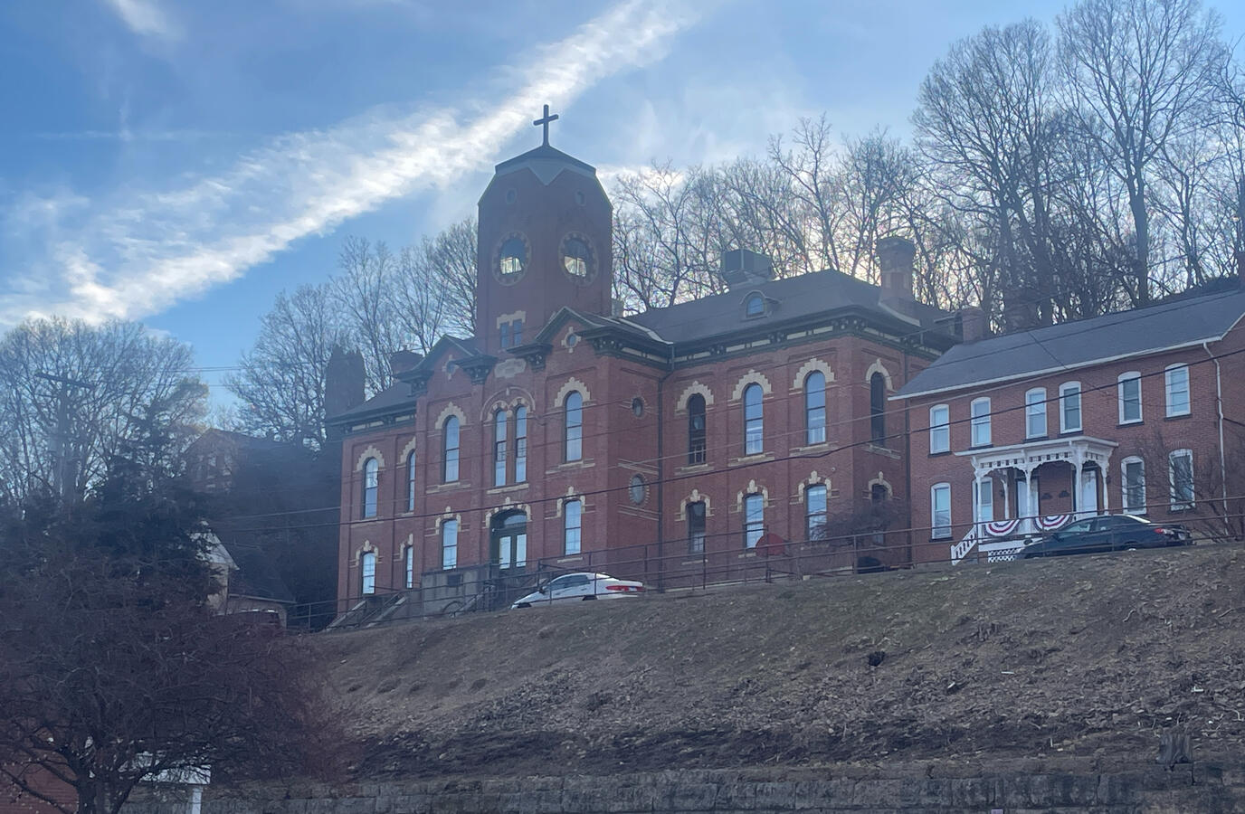 Historic 19th‑century brick building with a central tower and arched windows on a hilltop in Galena, Illinois, photographed in winter with leafless trees, parked cars, and a nearby brick home under a partly cloudy sky.