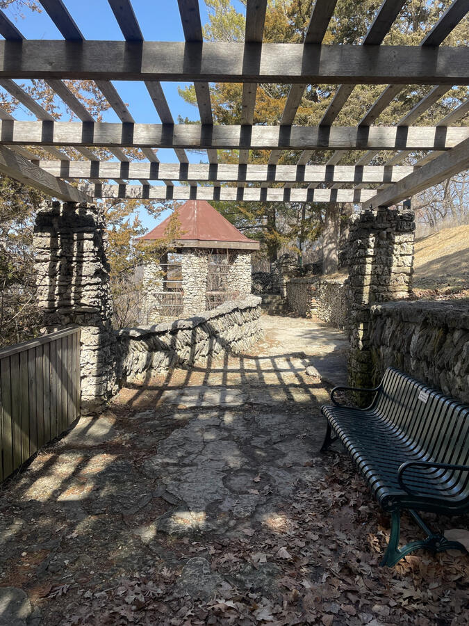 Stone pergola casting patterned shadows along a walkway in Phelps Park in Decorah.