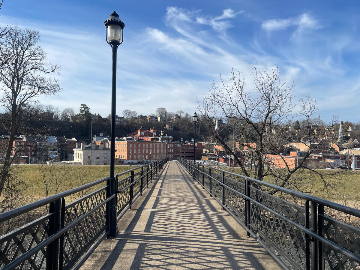 A pedestrian bridge provides open views across the quiet river corridor. View from the pedestrian bridge in Grant Park, Galena, Illinois, looking toward the historic downtown with brick buildings, church steeples, bare trees, and patterned shadows cast by the bridge’s metal railings on a clear early‑spring day.