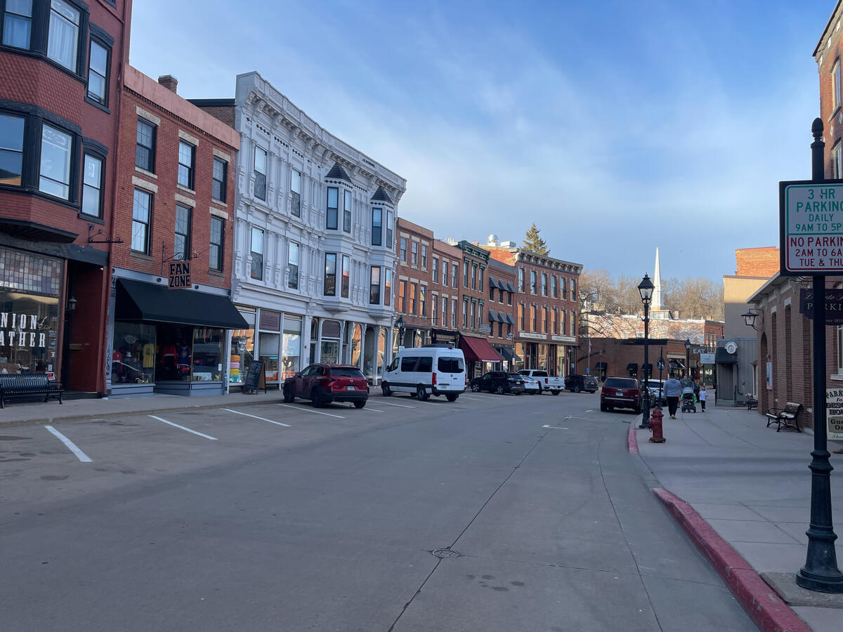 Narrow storefronts and cast-iron columns recall the era when Galena thrived as a regional trade center. Historic brick storefronts along Main Street in downtown Galena, Illinois, featuring shops like Fan Zone and Union Leather, parked cars, pedestrians, and a street sign indicating local parking hours.