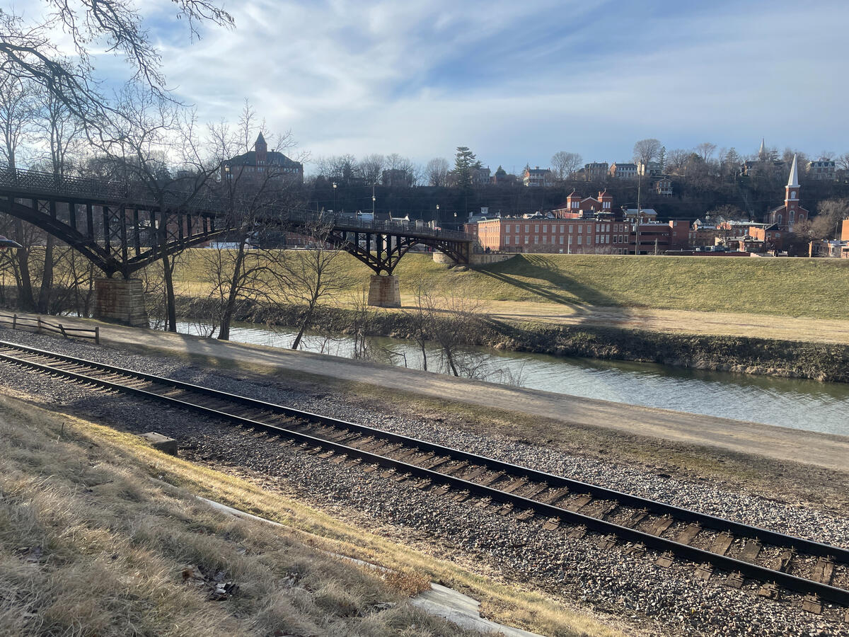 Rail bridges crossing the Galena River recall the transportation routes that once carried Galena’s trade outward. View from Grant Park in Galena, Illinois, showing railroad tracks beside the Galena River, a metal pedestrian bridge with stone supports, and historic brick buildings rising on the hillside under a partly cloudy sky.