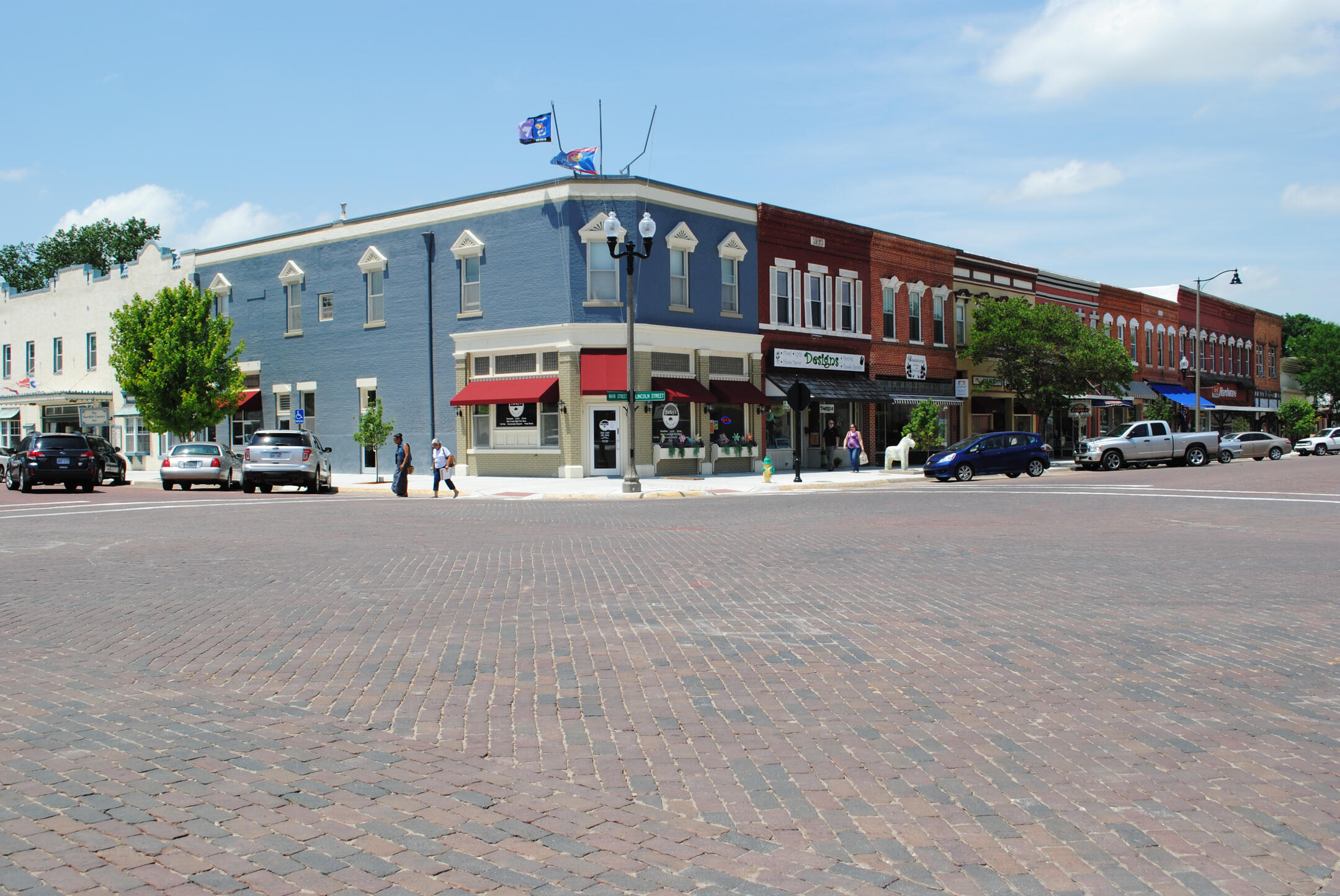 lindsborg-kansas-downtown-intersection-brick-street A brick‑paved downtown intersection in Lindsborg, Kansas, with historic storefronts, red awnings, parked cars, and people walking along the sidewalk on a sunny day.