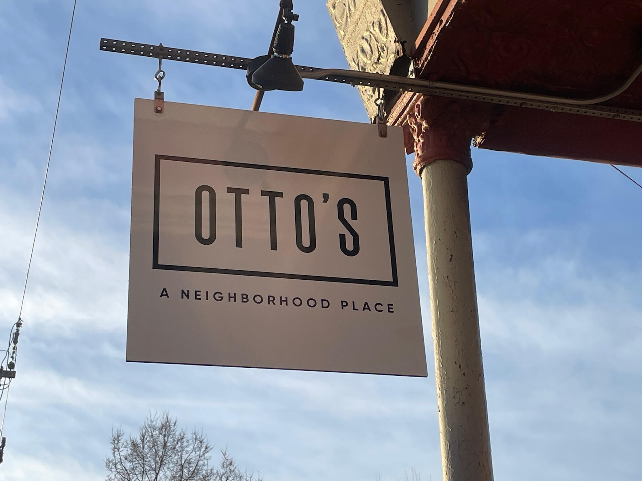otto's-sign-galena-illinois Outdoor hanging sign for Otto's Place restaurant in Galena, Illinois, featuring the minimalist black-and-white logo with the tagline "A neighborhood place" against a blue sky.