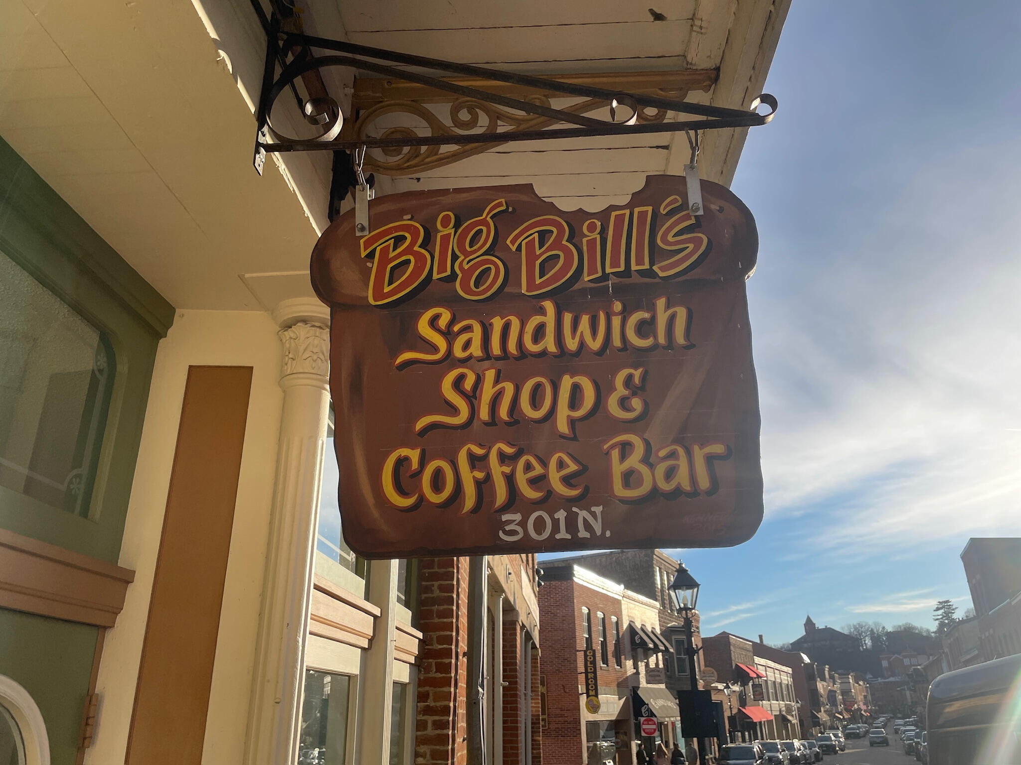 Exterior wooden hanging sign for Big Bill's Sandwich Shop & Coffee Bar located at 301 N. Main St. in Galena, Illinois, featuring yellow, white, and red hand-painted lettering on a brown bread-shaped board.