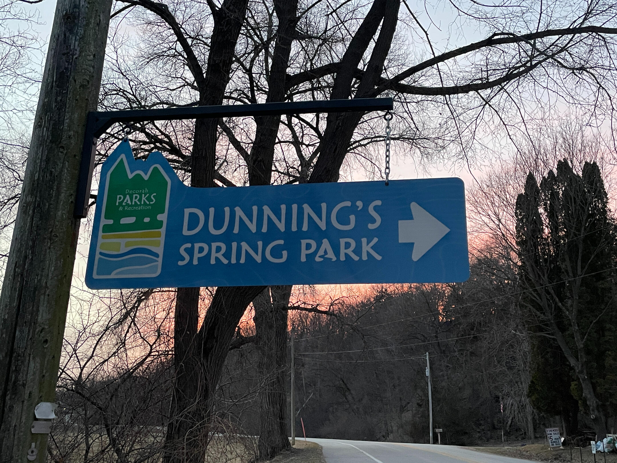 Blue Dunning’s Spring Park sign with an arrow pointing right beside a wooded roadside in Decorah.