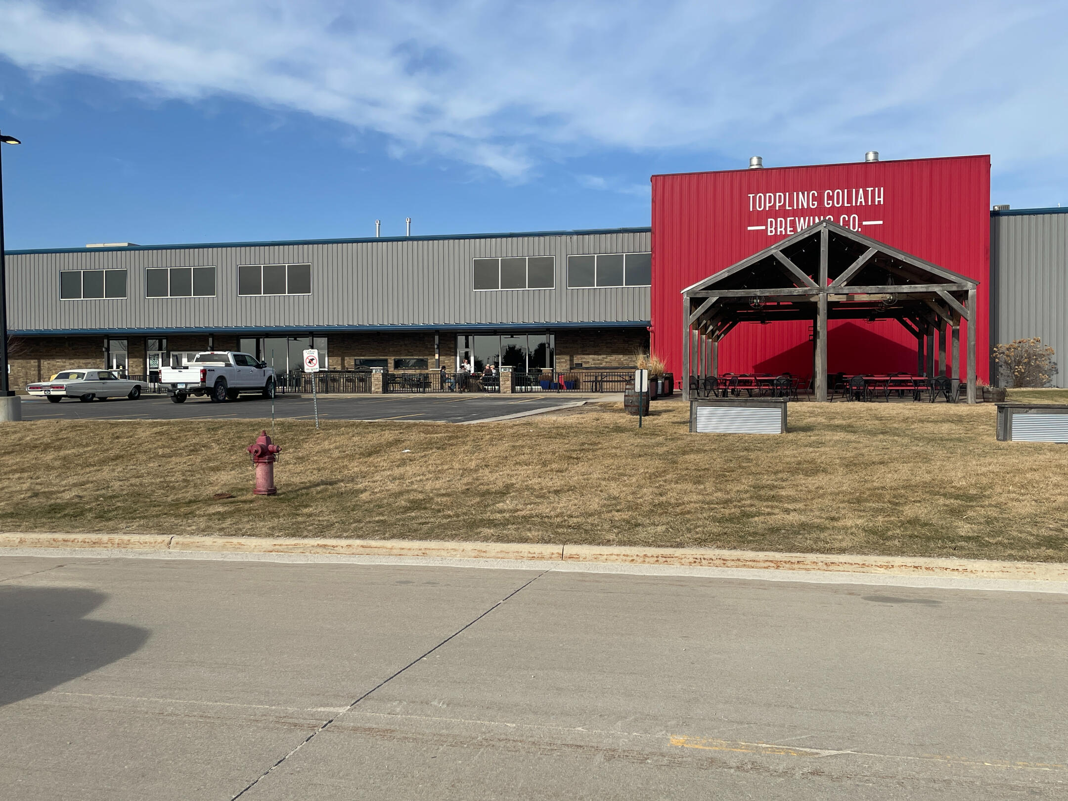 Industrial‑style Toppling Goliath Brewing Co. building with red siding, pavilion entrance, and parked vehicles outside Decorah.