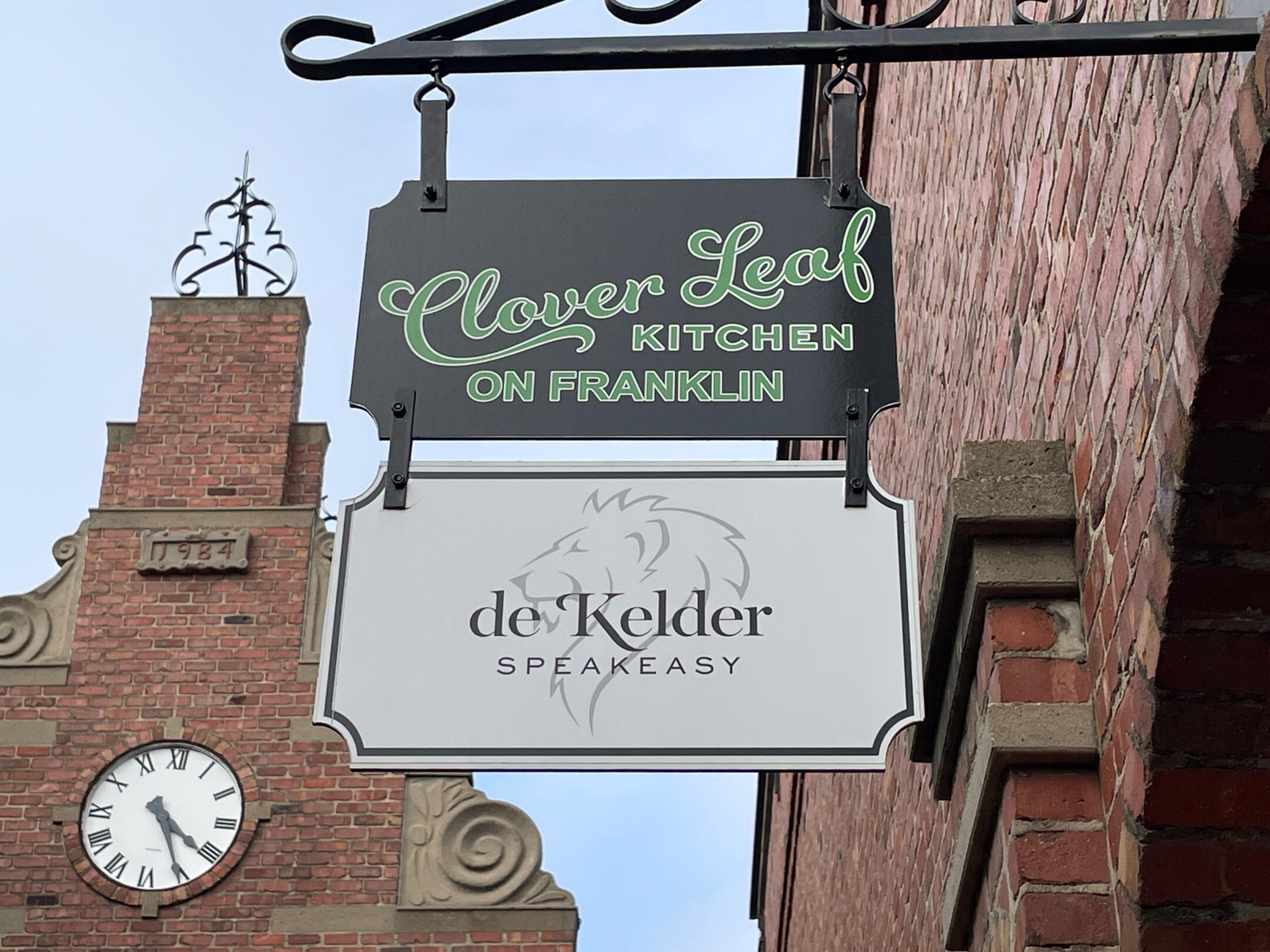 Hanging signs for Clover Leaf Kitchen and De Kelder Speakeasy mounted on a brick building in downtown Pella.