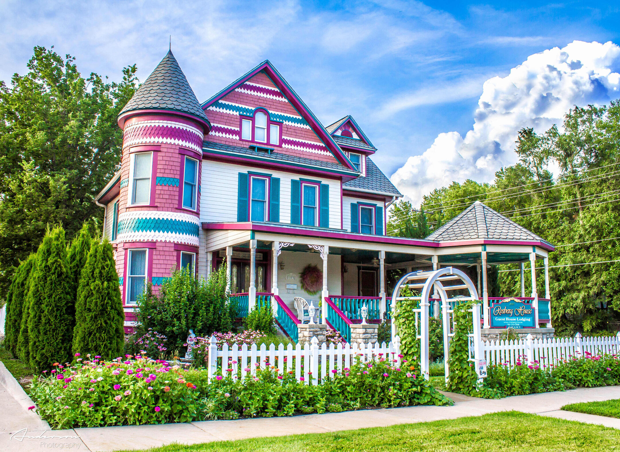 lindsborg-kansas-rosberg-house-victorian-guesthouse The Rosberg House in Lindsborg, Kansas, a brightly painted Victorian home with a turret, wraparound porch, white picket fence, and blooming garden.